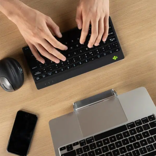 Healthy Home & Office - A person's hands are typing on a compact, black R-Go Break Keyboard placed on a wooden desk surface. A laptop, smartphone, and a computer mouse are also visible on the desk.