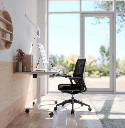 Black office chair next to a talent 500 height adjustable desk with wheels, in a room with wooden floor and big windows.