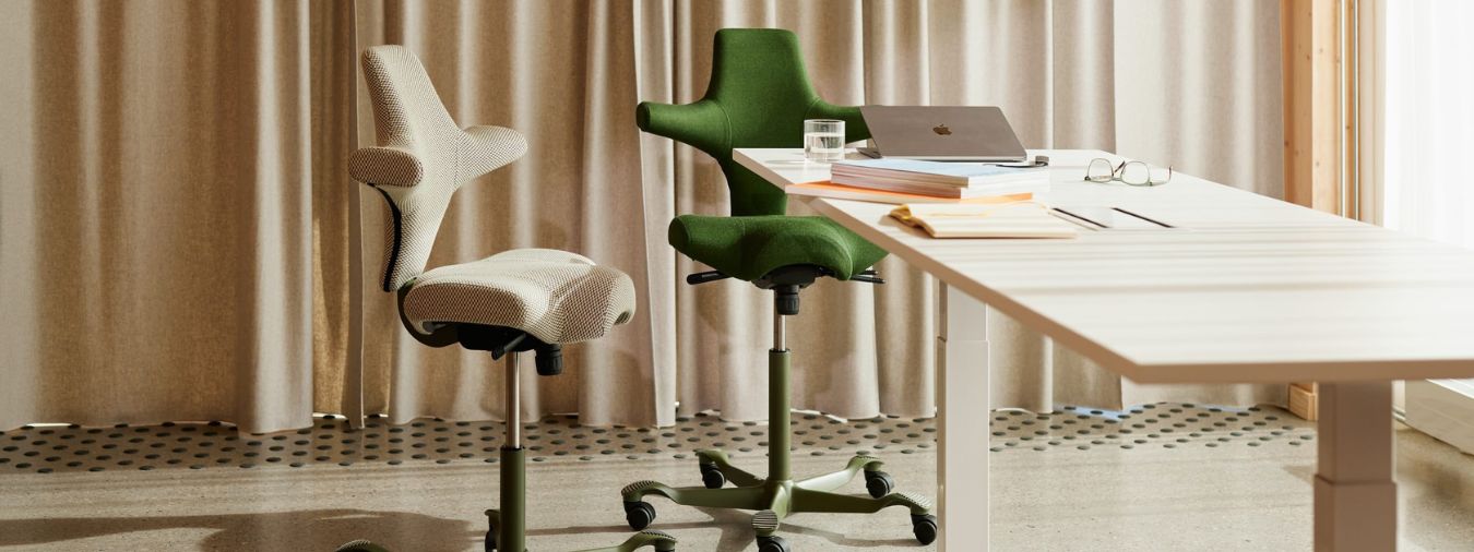 Two Capisco saddles chairs, one green and another beige, next to a height adjustable desk.
