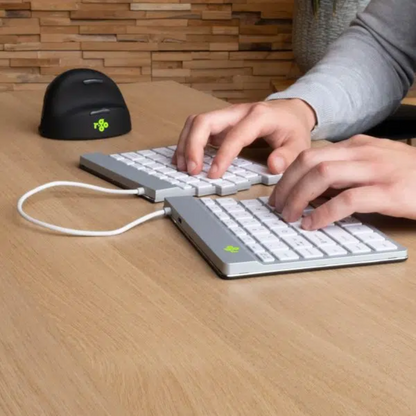 Healthy Home & Office - A person's hands typing on a split ergonomic keyboard with a white cable, positioned on a wooden desk surface alongside a black computer mouse.