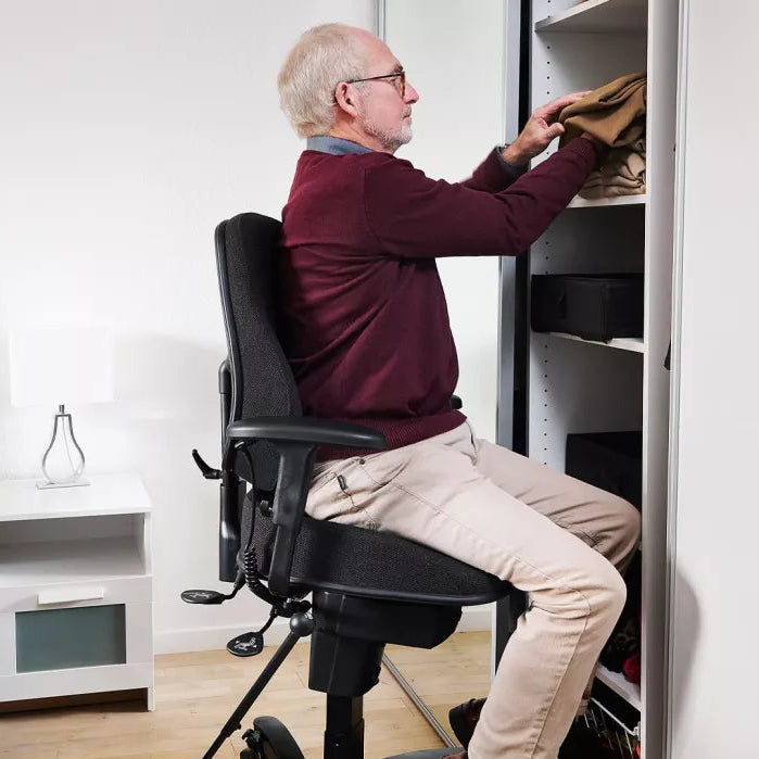 An older man with white hair and glasses sits in a black office chair, reaching into a tall white wardrobe or closet shelf to organize clothes. The scene is set in a modern bedroom with a white nightstand and lamp.