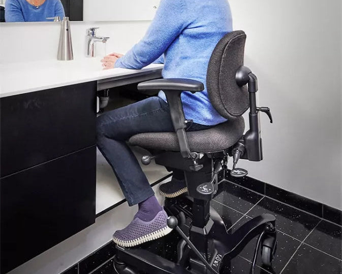 A woman sits on a black ergonomic office chair in a modern bathroom or office space, with a mirror showing her reflection behind a white sink and counter.